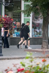 Фото тату на ноге девушки, идущей по улице. Рисунок выполнен в стиле стрит-арт, с использованием черных линий и небольших цветовых акцентов. Девушка одета в шорты и сетчатые колготки.
