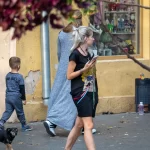 Фотография молодой девушки с короткими светлыми волосами, идущей по улице в яркой футболке и смотрящей в телефон. На руке видна татуировка.