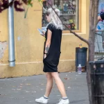 Фотография молодой женщины с короткими светлыми волосами, идущей по улице в черном платье и смотрящей в телефон. На плече видна татуировка.
