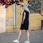 Фотография молодой женщины с короткими светлыми волосами, идущей по улице в черном платье и смотрящей в телефон. На плече видна татуировка.