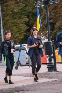 Фотография девушки с татуировкой на руке, идущей по улице на фоне украинского флага и городского пейзажа.
