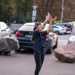 Фотография девушки с татуировкой на руке, делающей селфи на фоне городского пейзажа с автомобилями и пешеходами.
