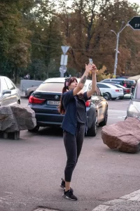 Фотография девушки с татуировкой на руке, делающей селфи на фоне городского пейзажа с автомобилями и пешеходами.