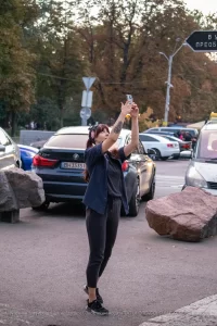 Фотография девушки с татуировкой на руке, делающей селфи на фоне городского пейзажа с автомобилями и пешеходами.