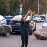 Фотография девушки с татуировкой на руке, делающей селфи на фоне городского пейзажа с автомобилями и пешеходами.