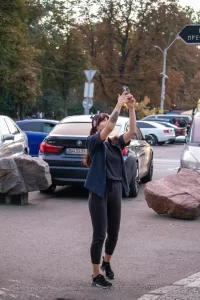 Фотография девушки с татуировкой на руке, делающей селфи на фоне городского пейзажа с автомобилями и пешеходами.