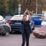 Фотография девушки с татуировкой на руке, делающей селфи на фоне городского пейзажа с автомобилями и пешеходами.
