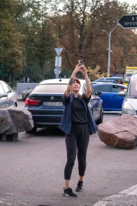 Фотография девушки с татуировкой на руке, делающей селфи на фоне городского пейзажа с автомобилями и пешеходами.