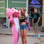 Фотография уличной татуировки: девушка радостно улыбается, позируя рядом с человеком в костюме Розовой Пантеры на городской улице.
