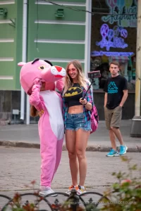 Фотография уличной татуировки: девушка радостно улыбается, позируя рядом с человеком в костюме Розовой Пантеры на городской улице.