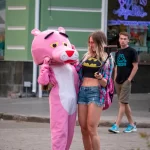 Фотография уличной татуировки: девушка в костюме Розовой Пантеры позирует с человеком, делающим селфи на фоне городской улицы.