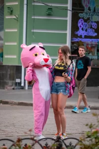 Фотография уличной татуировки: девушка в костюме Розовой Пантеры позирует с человеком, делающим селфи на фоне городской улицы.
