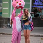 Фотография девушки в костюме розовой пантеры на улице. Девушка в розовом костюме и с головой в виде розовой пантеры, позирует на фоне городской среды.