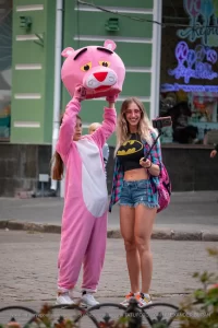 Фотография девушки в костюме розовой пантеры на улице. Девушка в розовом костюме и с головой в виде розовой пантеры, позирует на фоне городской среды.