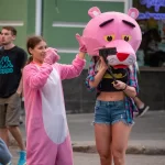 Фотография девушки в костюме розовой пантеры на улице. Девушка в розовом костюме и с головой в виде розовой пантеры, позирует на фоне городской среды.