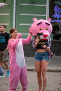 Фотография девушки в костюме розовой пантеры на улице. Девушка в розовом костюме и с головой в виде розовой пантеры, позирует на фоне городской среды.
