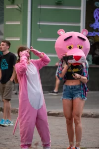 Фотография девушки в костюме розовой пантеры на улице. Девушка в розовом костюме и с головой в виде розовой пантеры, позирует на фоне городской среды.