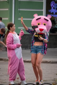 Фотография девушки в костюме розовой пантеры на улице. Девушка в розовом костюме и с головой в виде розовой пантеры, позирует на фоне городской среды.