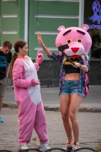 Фотография девушки в костюме розовой пантеры на улице. Девушка в розовом костюме и с головой в виде розовой пантеры, позирует на фоне городской среды.