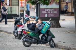 Фотография уличного трюка на мотоцикле. Человек выполняет сложный акробатический элемент, сидя на мотоцикле. На заднем плане видны здания и рекламные вывески.
