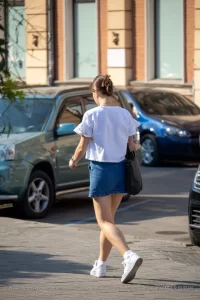 Фото тату: девушка идет по улице, демонстрируя татуировку птицы на плече. Рисунок выполнен в черных тонах, реалистичный стиль. Вид сзади.