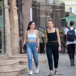 Фото тату: девушка с татуировкой птицы на плече идет по улице. Рисунок выполнен в черных тонах, реалистичный стиль. Городской фон.