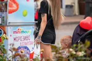 Фотография девушки с татуировкой на плече, стоящей у киоска с мороженым. Видна часть татуировки, предположительно, в виде цветка.