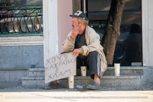Фотография уличной татуировки: мужчина с татуировкой Бэтмена на руке сидит на ступеньках с табличкой.