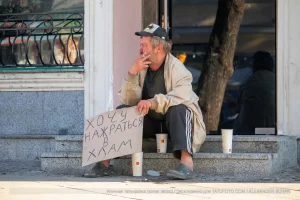 Фотография уличной татуировки: мужчина с татуировкой Бэтмена на руке сидит на ступеньках с табличкой.