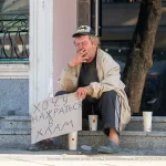 Фотография уличной татуировки: мужчина с татуировкой Бэтмена на руке сидит на ступеньках с табличкой.