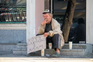 Фотография уличной татуировки: мужчина с татуировкой Бэтмена на руке сидит на ступеньках с табличкой.