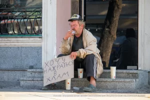 Фотография уличной татуировки: мужчина с татуировкой Бэтмена на руке сидит на ступеньках с табличкой.