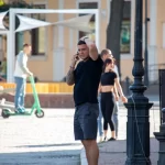 Фотография мужчины с татуировкой на предплечье, разговаривающего по телефону на улице. Он одет в шорты и футболку.