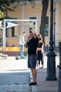 Фотография мужчины с татуировкой на предплечье, разговаривающего по телефону на фоне городской архитектуры. Татуировка выполнена в темных тонах.