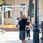 Фото тату: Мужчина с татуировкой на руке, говорящий по телефону на улице. Уличная фотография, демонстрирующая детализированный рисунок татуировки и повседневную сцену.
