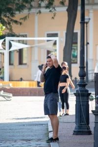 Фото тату: Мужчина с татуировкой на руке, говорящий по телефону на улице. Уличная фотография, демонстрирующая детализированный рисунок татуировки и повседневную сцену.