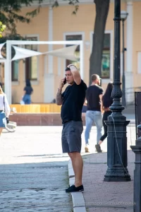 Фото тату: Мужчина с татуировкой на руке, говорящий по телефону на улице. Уличная фотография, демонстрирующая детализированный рисунок татуировки и повседневную сцену.