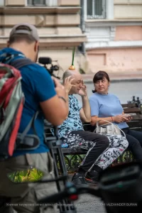 Фото тату: Мужчина с татуировкой на плече и луковицей на голове, сидящий на скамейке с женщиной. Уличная фотография, демонстрирующая необычный образ и татуировку.