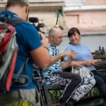 Фото тату: Мужчина с татуировкой на плече и луковицей на голове, сидящий на скамейке с женщиной. Уличная фотография, демонстрирующая необычный образ и татуировку.