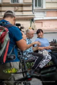 Фото тату: Мужчина с татуировкой на плече и луковицей на голове, сидящий на скамейке с женщиной. Уличная фотография, демонстрирующая необычный образ и татуировку.