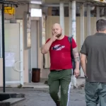 Фото тату: Мужчина с татуировкой ворона на предплечье, курящий сигарету и смотрящий вдаль. Уличная фотография, демонстрирующая детализированный рисунок татуировки и атмосферу раздумий.