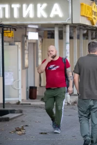 Фото тату: Мужчина с татуировкой ворона на предплечье, курящий сигарету и смотрящий вдаль. Уличная фотография, демонстрирующая детализированный рисунок татуировки и атмосферу раздумий.