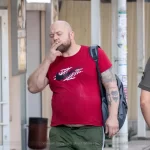 Фото тату: Мужчина с татуировкой ворона на предплечье, курящий сигарету. Уличная фотография, демонстрирующая детализированный рисунок татуировки и повседневную сцену.