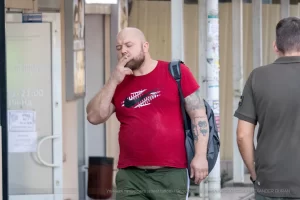 Фото тату: Мужчина с татуировкой ворона на предплечье, курящий сигарету. Уличная фотография, демонстрирующая детализированный рисунок татуировки и повседневную сцену.