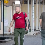 Фото тату: Мужчина с татуировкой черного ворона на предплечье, летящего среди красных брызг краски. Уличная фотография, демонстрирующая детализированный рисунок татуировки.