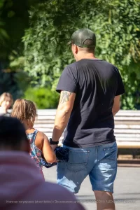 Фото тату птицы на руке мужчины, который ведет за руку маленькую девочку. Рисунок выполнен в черных тонах, с детальной проработкой.