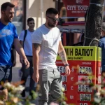 Фото мужчины с татуировками на руке и шее, стоящего на фоне уличного обмена валют, демонстрирующее современный стиль татуировки.