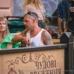 Фотография мужчины в кепке и темной одежде с татуировкой волка на руке, сидящего за столиком в кафе.