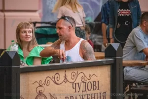 Фотография мужчины в кепке и темной одежде с татуировкой волка на руке, сидящего за столиком в кафе.