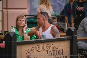 Крупный план руки мужчины с яркой татуировкой, сидящего в уличном кафе. Видны детали рисунка и окружающая обстановка.